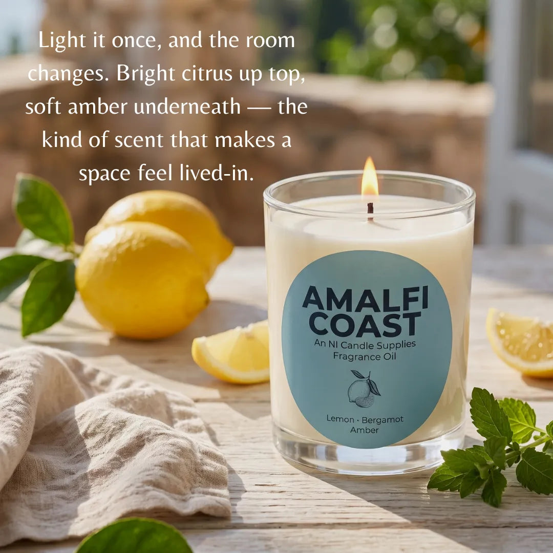 A lit candle in a clear glass jar sits on a wooden table with lemon slices, mint leaves, and a beige cloth. The candle label reads AMALFI COAST and specifies Lemon - Bergamot and Amber scent.