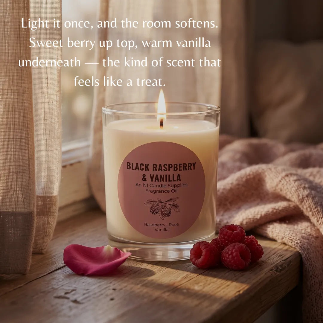 A lit scented candle in a clear glass jar sits on a wooden windowsill with a warm cozy setting. The candle has a cream-colored wax and a dusty rose circular label that reads 'BLACK RASPBERRY & VANILLA'.