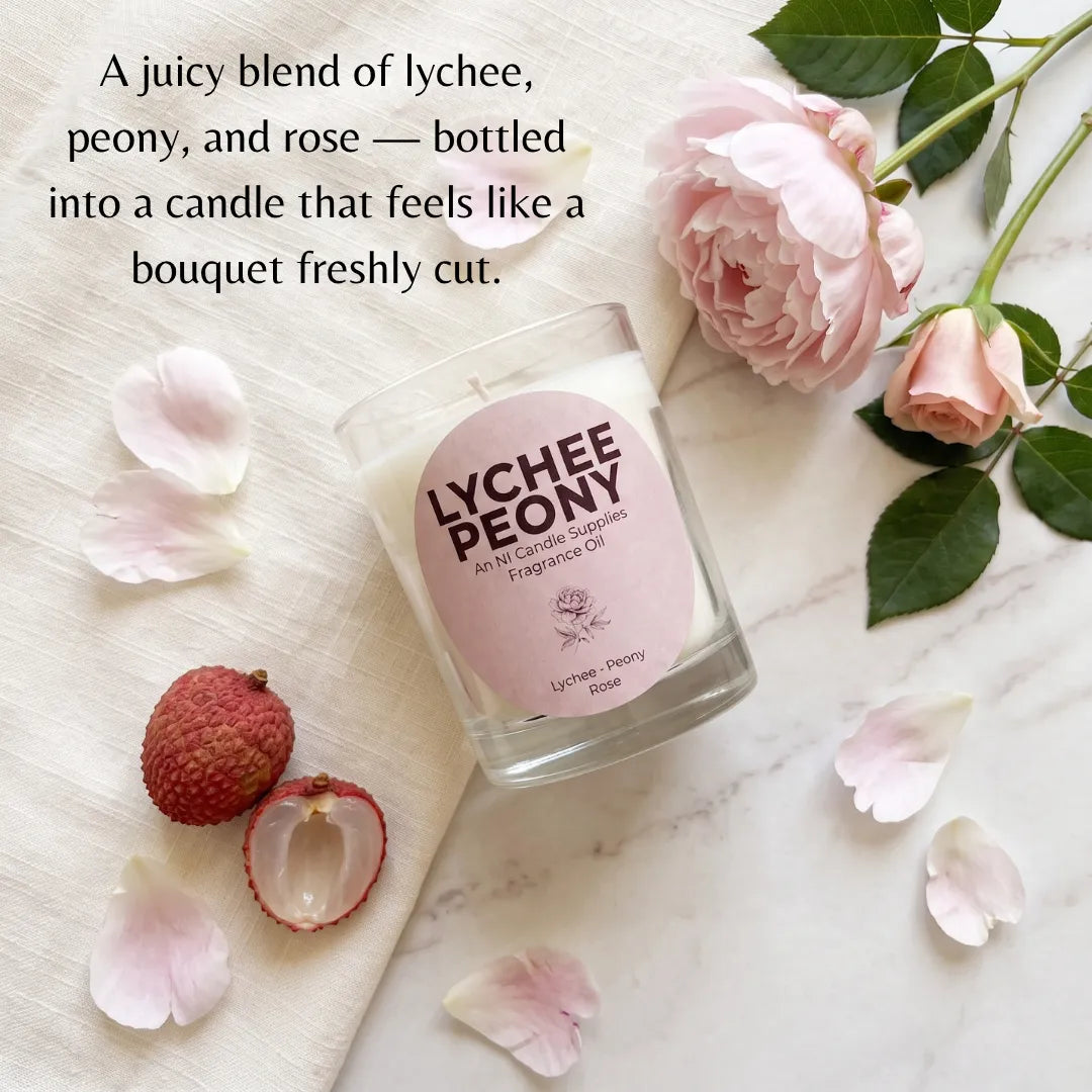 A clear glass candle jar labeled 'LYCHEE PEONY' sits on a light marble surface with pink roses, petals, and lychee fruit around it. Text at the top reads, 'A juicy blend of lychee, peony, and rose — bottled into a candle that feels like a bouquet freshly cut.
