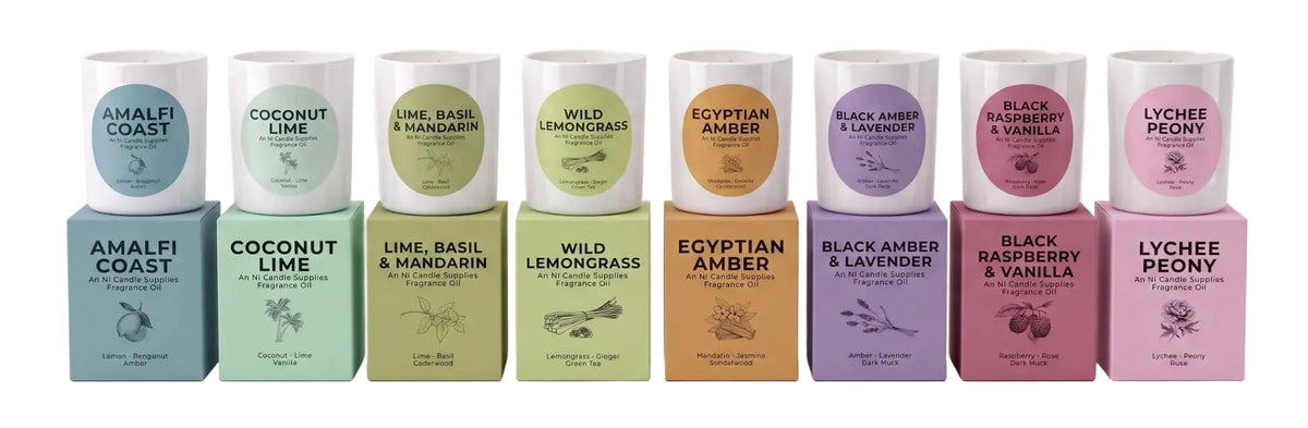 A row of eight candles with matching colored boxes, each labeled with a different scent name including Amalfi Coast, Coconut Lime, Lime & Basil & Mandarin, Wild Lemongrass, Egyptian Amber, Black Amber & Lavender, Black Raspberry & Vanilla, and Lychee Peony, arranged on a plain white background.