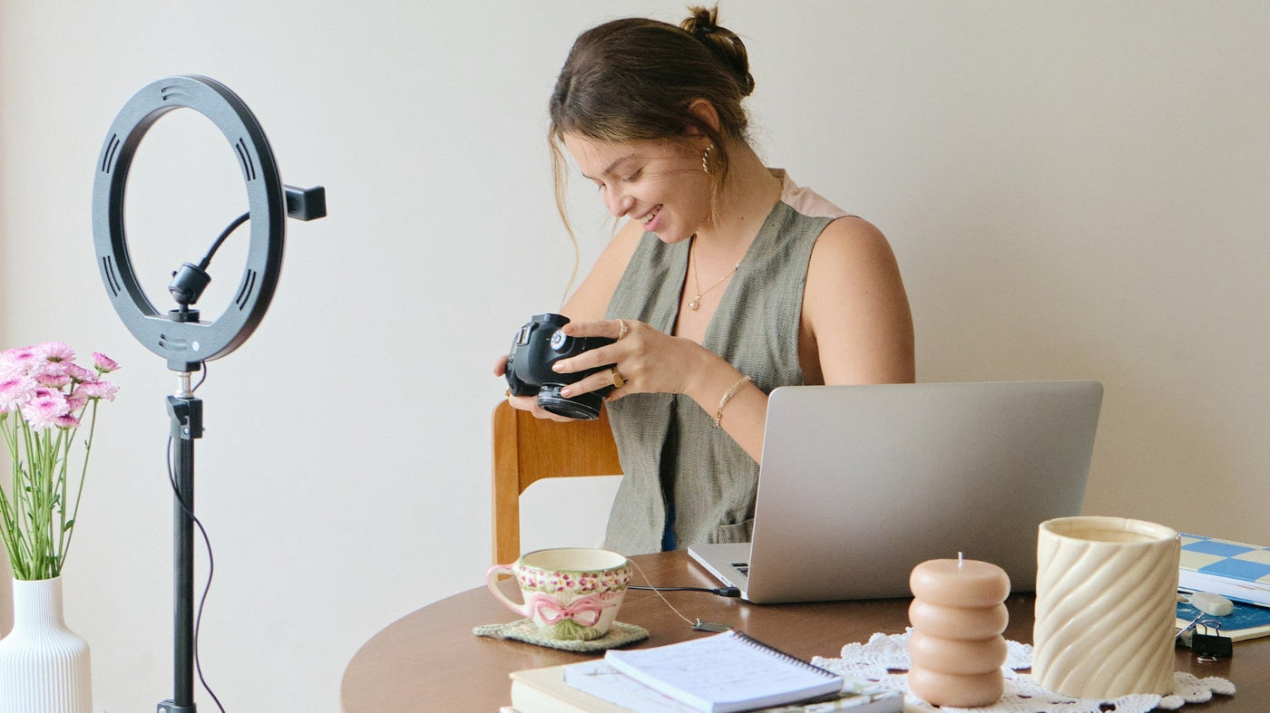 Woman sitting at a table with a laptop, camera, and ring light in a home setting.