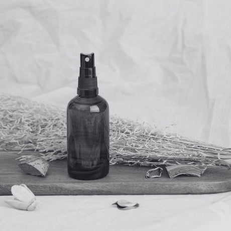 Room Spray Supplies - Dark glass spray bottle on wooden board with dried botanicals and potpourri, styled in a neutral, minimalist setting.