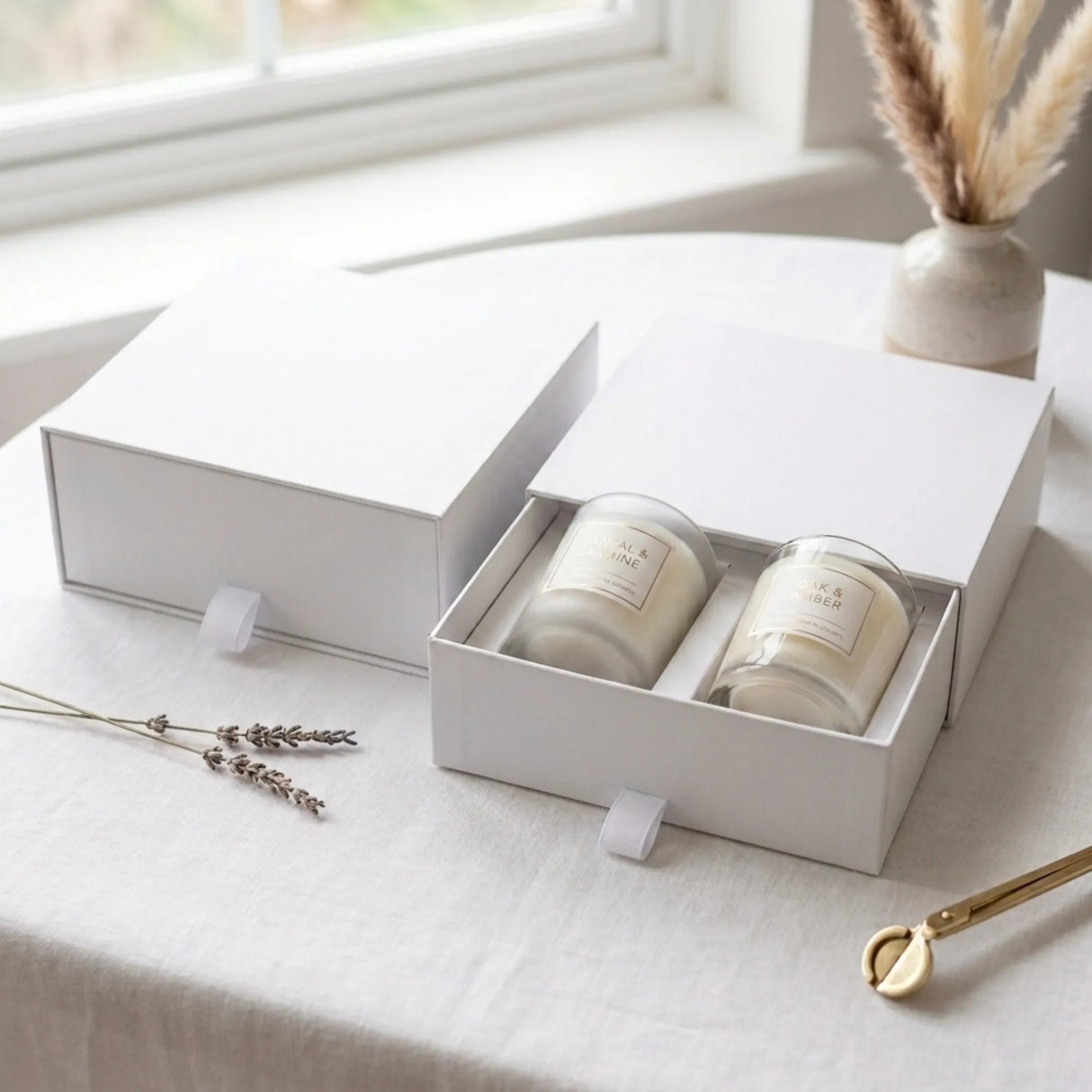 Two white jar candles in a white gift box set on a light tablecloth near a window, with dried grass and a gold candle tongs prop nearby.