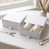 Two white jar candles in a white gift box set on a light tablecloth near a window, with dried grass and a gold candle tongs prop nearby.