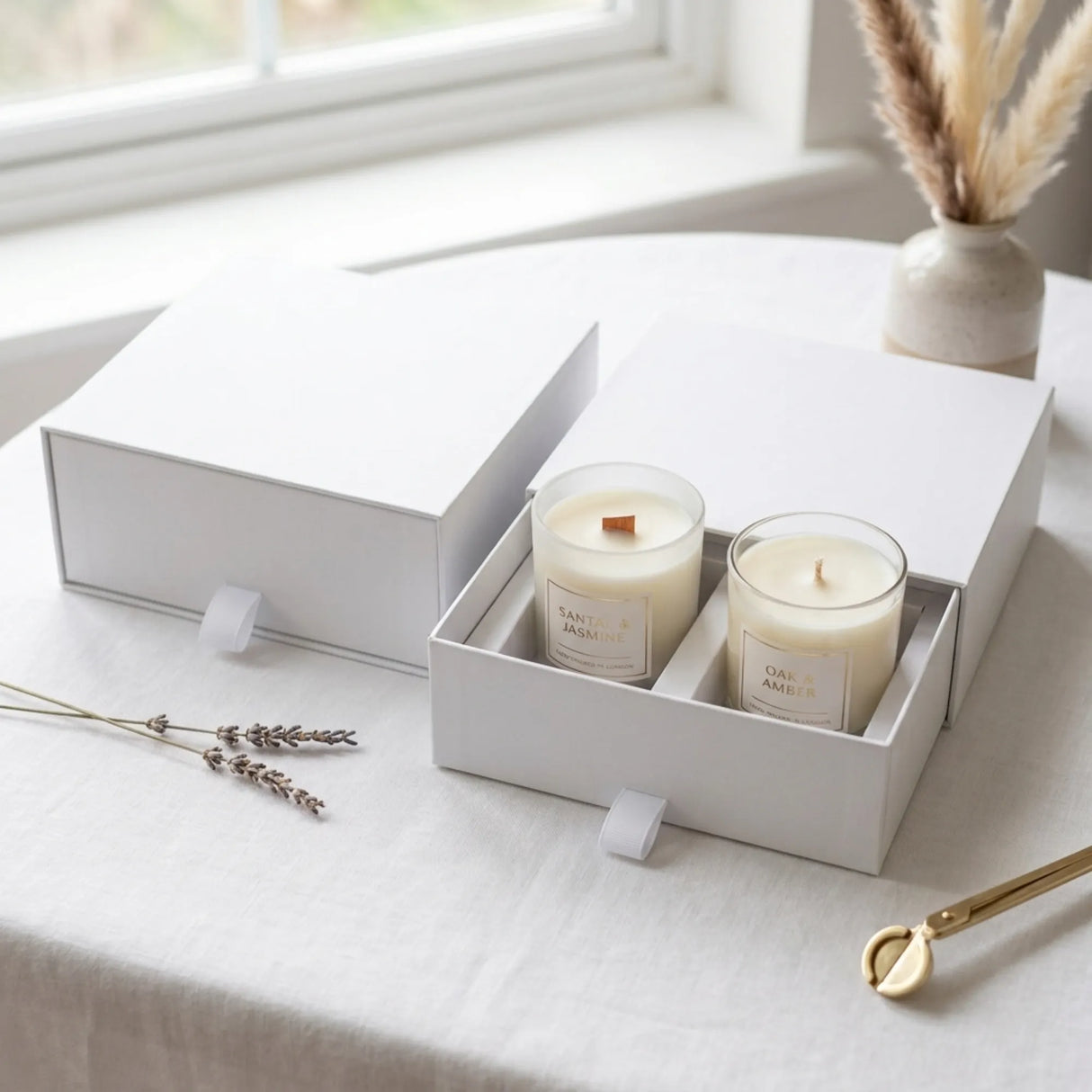 Two white soy candles in clear glass jars inside a white gift box, set on a light tablecloth near a window with dried lavender and a vase in the background.