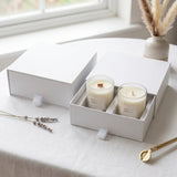 Two white soy candles in clear glass jars inside a white gift box, set on a light tablecloth near a window with dried lavender and a vase in the background.