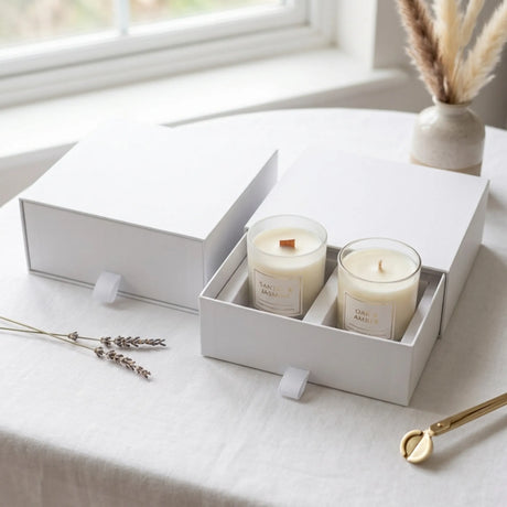 Two white soy candles in clear glass jars inside a white gift box, set on a light tablecloth near a window with dried lavender and a vase in the background.