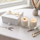 Two lit scented candles in clear glass jars beside a white gift box with a cream ribbon and gold tag, styled on a white tablecloth near a window with dried grass and lavender sprigs.