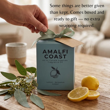 A hand holds a sage green ribbon bow tied to a light blue box labeled AMALFI COAST Fragrance Oil. The box sits on a wooden table with lemon slices, eucalyptus leaves, and a blurred indoor background.