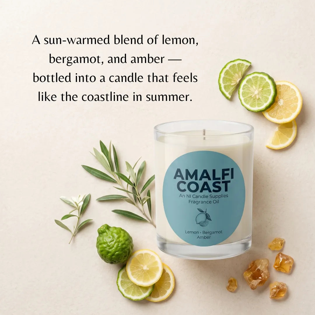 A clear glass candle jar labeled AMALFI COAST sits on a light beige background with lemon slices, lime wedges, green leaves, and amber stones. Text above reads: 'A sun-warmed blend of lemon, bergamot, and amber — bottled into a candle that feels like the coastline in summer.