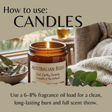 A lit amber glass candle jar labeled 'AUSTRALIAN BUSH' sits on a table in a cozy indoor setting with a blanket, leaves, and a cup in the background. Large text at the top reads 'How to use: CANDLES' and text at the bottom explains fragrance oil loading for a clean burn.