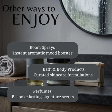 Promotional graphic for a bathroom or spa wellness product collection, showing room sprays, bath & body products, and perfumes on a dark countertop with overlaid text labels. The scene includes a towel, a round mirror with raindrops, and a gray wall.