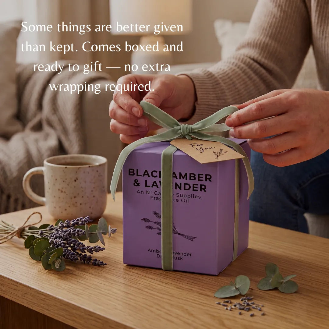 A person's hands tie a sage green ribbon on a purple gift box labeled 'Black &amp; White' and 'Blackambers & Lavender'. The box sits on a wooden table with a mug, lavender sprigs, and eucalyptus leaves in a cozy indoor setting.