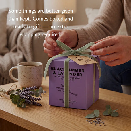 A person's hands tie a sage green ribbon on a purple gift box labeled 'Black &amp; White' and 'Blackambers & Lavender'. The box sits on a wooden table with a mug, lavender sprigs, and eucalyptus leaves in a cozy indoor setting.