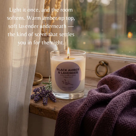 A lit glass jar candle labeled 'BLACK AMBER & LAVENDER' sits on a wooden windowsill with a draped purple knit blanket, lavender sprigs, sheer curtain, and soft outdoor light in the background. White text on the left describes the scent and mood.