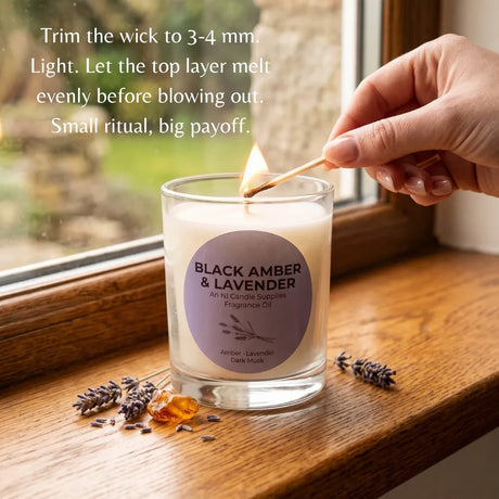 A hand lighting a glass jar candle with a match on a wooden windowsill. The candle label reads 'BLACK AMBER & LAVENDER' and 'An Ni Candle Supplies Fragrance Oil.