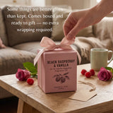 A pink square gift box tied with a pale pink ribbon sits on a wooden table in a cozy living room setting. A hand is reaching for the ribbon, and the box is labeled 'BLACK RASPBERRY & VANILLA' with raspberry and rose illustrations.