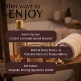 A promotional graphic for a spa or wellness product collection, showing several amber glass bottles and jars on a wooden shelf beside a rolled cream towel. Large overlaid text reads 'Other ways to ENJOY' with bullet points for 'Room Sprays', 'Bath & Body Products', and 'Perfumes'.