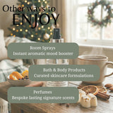A perfume or room spray bottle is centered on a wooden table with overlaid marketing text reading 'Other ways to ENJOY' and three callouts about room sprays, bath & body products, and perfumes. The scene includes a mug, dried orange slices, cinnamon, marshmallows, and a softly blurred holiday window background.