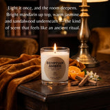 A lit scented candle in a clear glass jar sits on a wooden table in a warmly lit room, with a brass lamp, orange slices, cinnamon sticks, and white flowers around it. White text at the top reads, 'Light it once, and the room deepens.