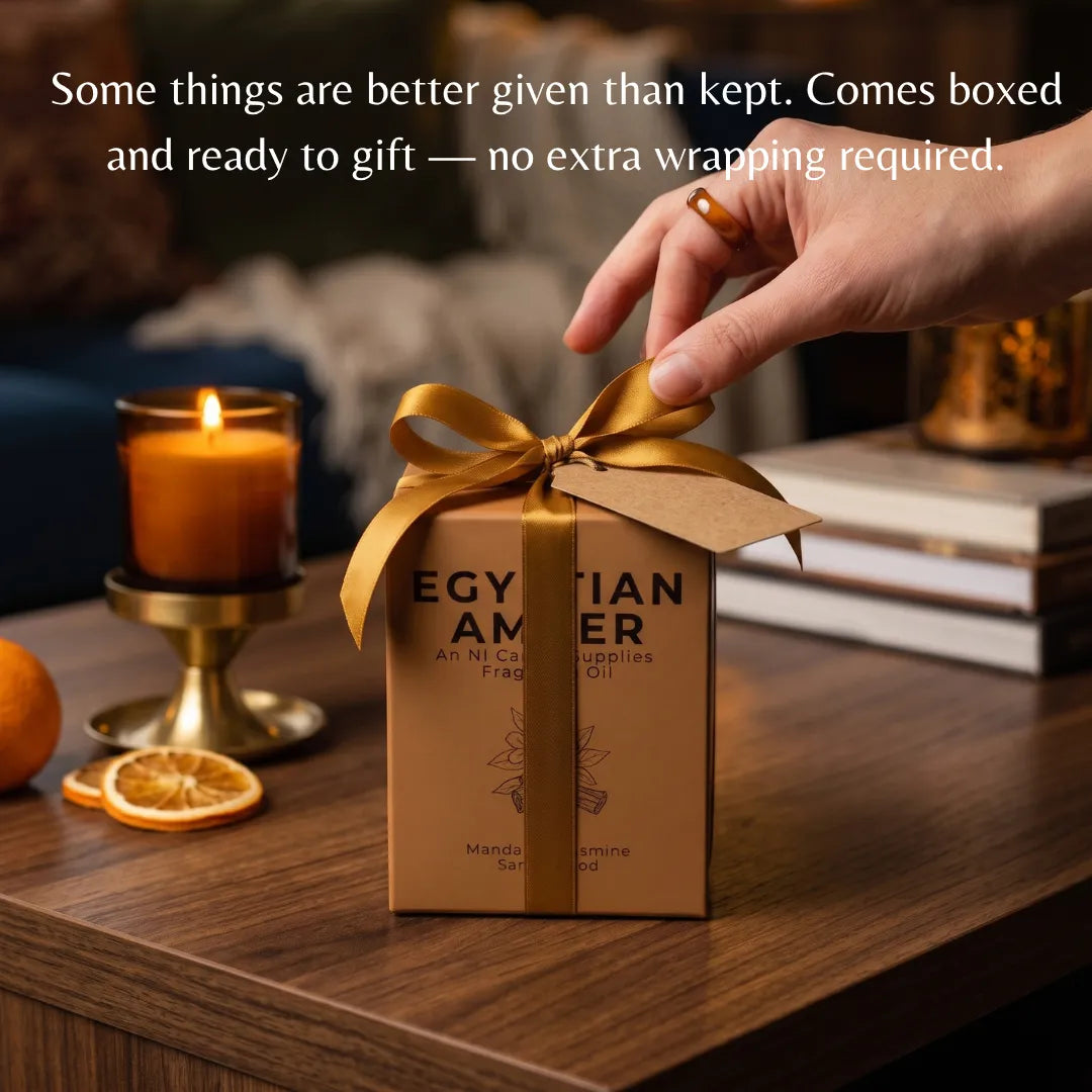 A hand places a gold ribbon on a brown gift box labeled 'EGYPTIAN AMBER' on a wooden table, with a lit candle, dried orange slices, and books in the background. White text at the top reads, 'Some things are better given than kept.