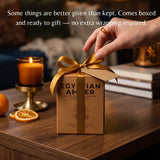 A hand places a gold ribbon on a brown gift box labeled 'EGYPTIAN AMBER' on a wooden table, with a lit candle, dried orange slices, and books in the background. White text at the top reads, 'Some things are better given than kept.