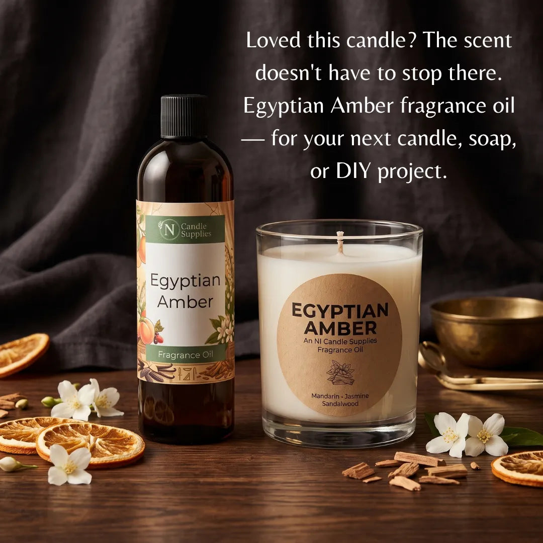 Egyptian Amber fragrance oil in a dark amber bottle next to a glass jar candle with a lit wick, both labeled Egyptian Amber, styled on a wooden surface with dried orange slices, white flowers, cinnamon sticks, and a gold bowl. Text on the image describes the scent and suggests using the fragrance oil for candles, soaps, or DIY projects.