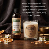 Egyptian Amber fragrance oil in a dark amber bottle next to a glass jar candle with a lit wick, both labeled Egyptian Amber, styled on a wooden surface with dried orange slices, white flowers, cinnamon sticks, and a gold bowl. Text on the image describes the scent and suggests using the fragrance oil for candles, soaps, or DIY projects.