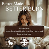 A woman sits on a sofa in a softly lit living room, holding a glass. Large text at the top reads 'Better Made BETTER BURN,' with a dark maroon banner below listing product benefits: 'Paraben-free, Natural soy-wax blend, Lead-free cotton wick, Long-lasting burn.