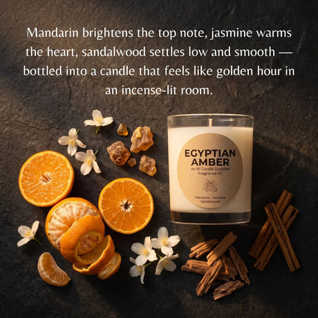 A glass jar candle labeled 'EGYPTIAN AMBER' sits on a dark textured surface with citrus slices, sandalwood pieces, cinnamon sticks, and small white flowers. A descriptive text overlay appears at the top describing the scent notes.