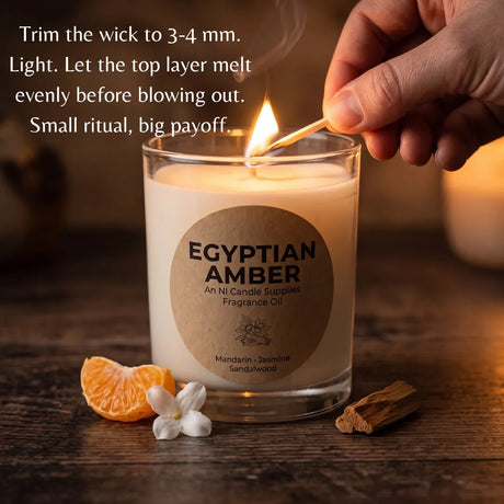 A hand holds a lit match next to an 'Egyptian Amber' scented candle in a clear glass jar on a wooden surface, with a candle wick being trimmed by another hand. Text on the top left explains the lighting process.