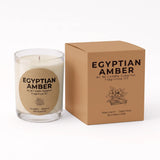 A clear glass jar candle with cream-colored wax and a round kraft paper label reading 'EGYPTIAN AMBER', shown next to its matching brown cardboard box on a white background.