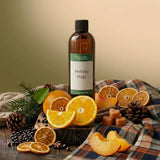 An amber glass bottle labeled 'Holiday Hugs' fragrance oil stands on a plaid fabric surface with orange slices, pinecones, dried orange slices, cinnamon sticks, and a few cubes around it. No person or animal is present, but this is a styled product scene rather than a plain studio cutout, so Product/Packaging fits better than Lifestyle.