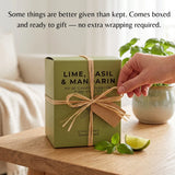 A sage green rectangular box labeled 'LIME, BASIL & MANGROU' sits on a wooden table tied with a tan ribbon. A hand reaches toward the box, with a potted plant and soft indoor background behind it.