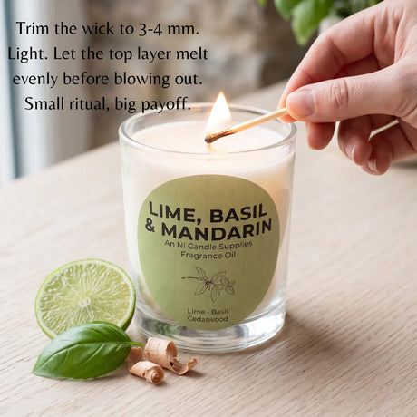 A lit scented candle in a clear glass jar sits on a light wood surface with a hand holding a match above the flame. A green circular label reads 'LIME, BASIL & MANDARIN' and 'An Ni Candle Supplies Fragrance Oil.