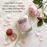 A clear glass candle jar labeled 'LYCHEE PEONY' sits on a light marble surface with pink roses, petals, and lychee fruit around it. Text at the top reads, 'A juicy blend of lychee, peony, and rose — bottled into a candle that feels like a bouquet freshly cut.