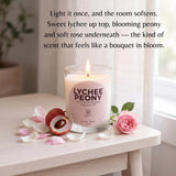 A lit glass jar candle labeled 'LYCHEE PEONY' sits on a light wood table with pink petals, a lychee fruit half, and a pink rose in a softly lit room. Text above describes the scent notes.