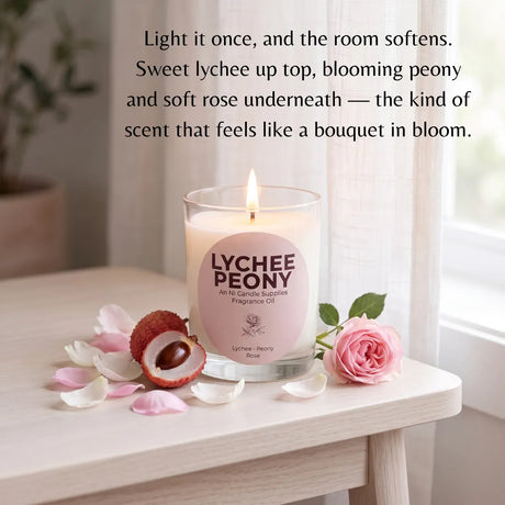 A lit glass jar candle labeled 'LYCHEE PEONY' sits on a light wood table with pink petals, a lychee fruit half, and a pink rose in a softly lit room. Text above describes the scent notes.