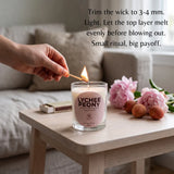 A hand holds a lit match over a glass jar candle on a light wood table in a softly lit room. The candle label reads 'LYCHEE PEONY' and includes scent and fragrance details.