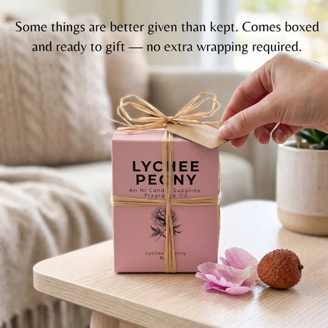 A pink Lychee Penny gift box tied with a natural jute ribbon and kraft paper tag, shown on a light wood table in a softly blurred indoor setting. A hand is touching the ribbon tag, and the text at the top reads, 'Some things are better given than kept.