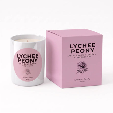 A white glass candle jar with a pink circular label next to its matching pink box, both printed with the text 'LYCHEE PEONY' and a rose illustration, on a clean white background.