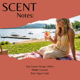 A promotional graphic for a fragrance or scent note, with a woman sitting outdoors by a lake holding a drink. Large coral text at the top reads 'SCENT Notes:' and the bottom text lists fragrance notes: 'Top: Lemon, Orange, Melon', 'Middle: Coconut', 'Base: Sugar Candy'.