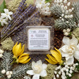 A square wax melt sample pack from Candle Society, labeled 'White Christmas', displayed in a clear plastic clamshell package. The package is beautifully arranged in a flat-lay on a rustic wooden background, surrounded by holiday-themed botanicals including purple lavender, white flowers, yellow tulips, and frosted green pine branches.