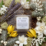 A square wax melt sample pack from Candle Society, labeled 'White Christmas', displayed in a clear plastic clamshell package. The package is beautifully arranged in a flat-lay on a rustic wooden background, surrounded by holiday-themed botanicals including purple lavender, white flowers, yellow tulips, and frosted green pine branches.