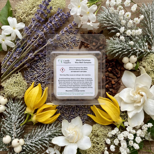 A square wax melt sample pack from Candle Society, labeled 'White Christmas', displayed in a clear plastic clamshell package. The package is beautifully arranged in a flat-lay on a rustic wooden background, surrounded by holiday-themed botanicals including purple lavender, white flowers, yellow tulips, and frosted green pine branches.