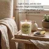 A lit candle in a clear glass jar sits on a stone tray beside a small dish of dried leaves and a lemon slice, with a linen chair and window in the background. The label reads “WILD LEMONGRASS” and “Masa Organic Lemongrass & Ginger Oil.