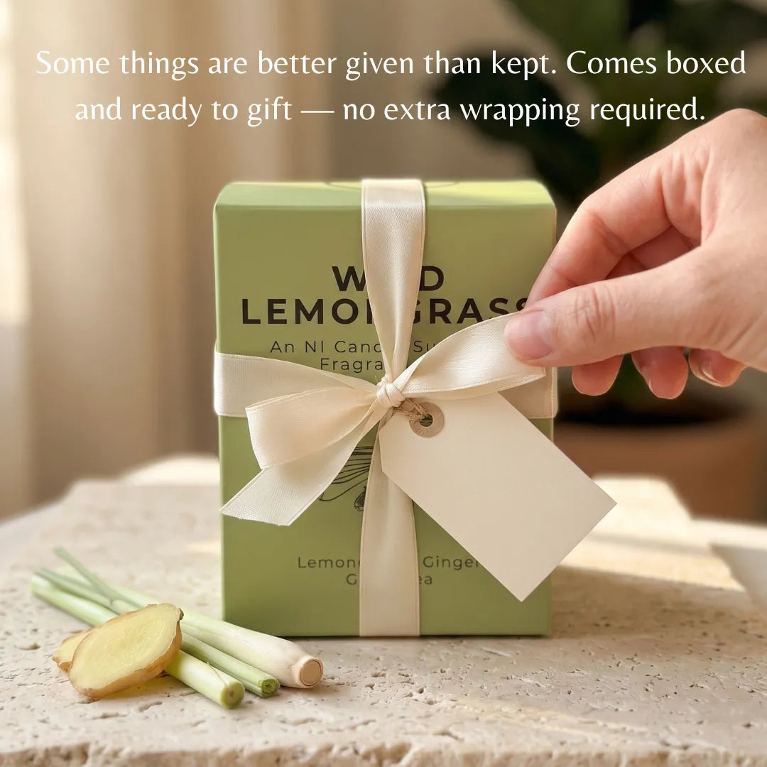 A light green rectangular candle box tied with a cream ribbon and bow, placed on a stone surface with lemongrass and ginger slices nearby. A hand is touching the ribbon.