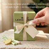 A light green rectangular candle box tied with a cream ribbon and bow, placed on a stone surface with lemongrass and ginger slices nearby. A hand is touching the ribbon.