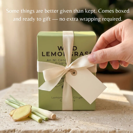 A light green rectangular candle box tied with a cream ribbon and bow, placed on a stone surface with lemongrass and ginger slices nearby. A hand is touching the ribbon.